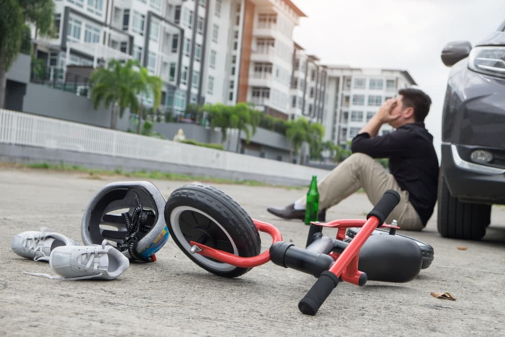 A scared and stressed, desperate drunken driver with a bottle of beer in front of an automobile crash involving a child's bike after a traffic accident on a city road.