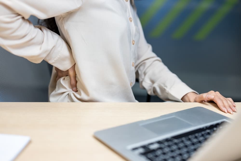 Office employee experiencing discomfort while seated at a desk, pressing their lower back to relieve strain or muscle pain.