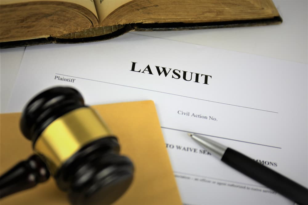 A close-up of legal documents titled "Lawsuit" lying on a wooden desk. The scene includes a gavel and block in the background, symbolizing the court's authority.