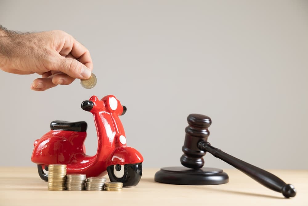 Close-up of a red motorbike-shaped piggy bank with a judge's gavel and money, set against a plain background.