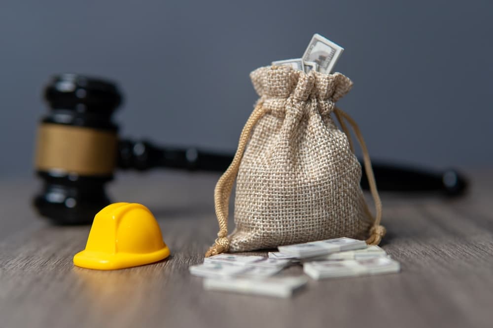 A closeup image of a money bag, hard hat, and gavel on a wooden table, symbolizing workers' compensation and labor law.