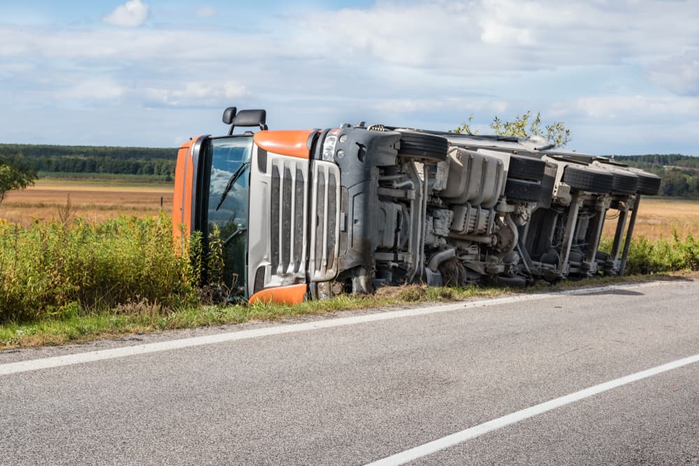 An overturned truck lies in a field beside the road, exposing its chassis.