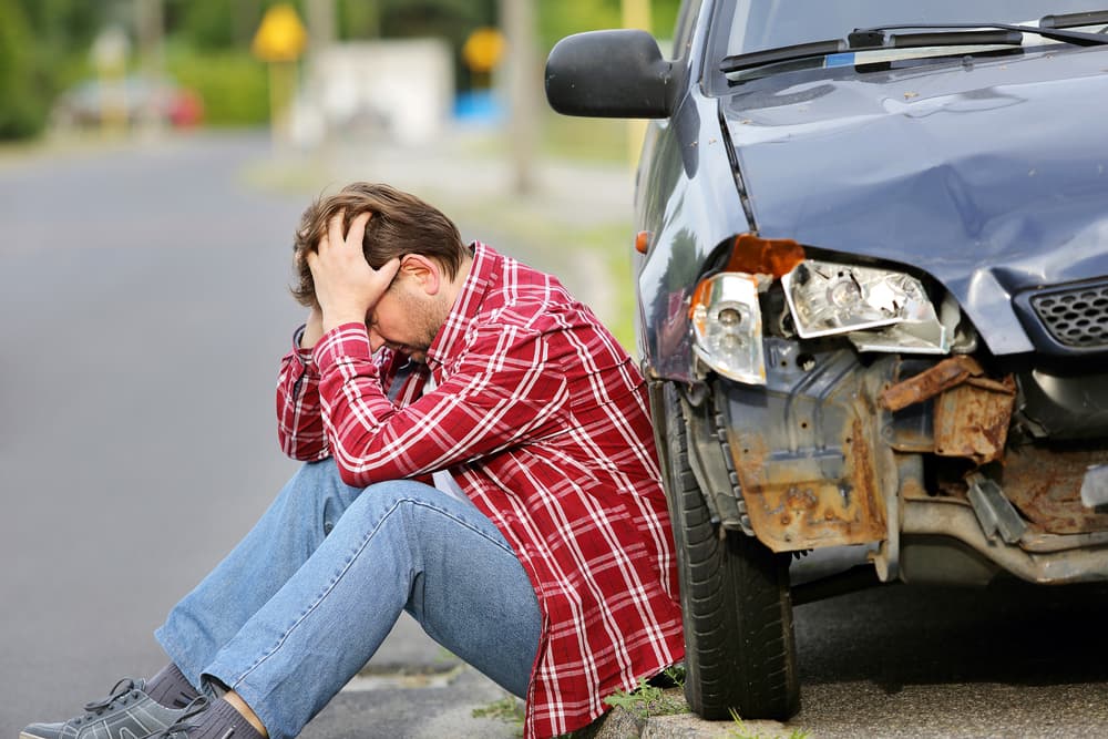 A man sitting on the ground near his car after an accident, looking distressed. The car has visible damage, and debris is scattered around. 