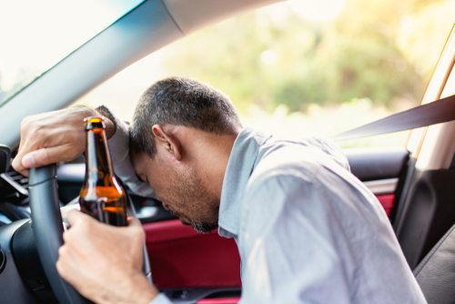 Man slumped over a steering wheel holding a beer bottle, illustrating the dangers and consequences of drunk driving.