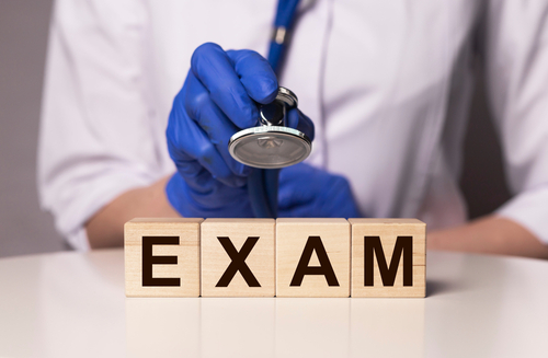 Doctor in gloves holding a stethoscope above wooden blocks spelling "EXAM," symbolizing a medical examination.