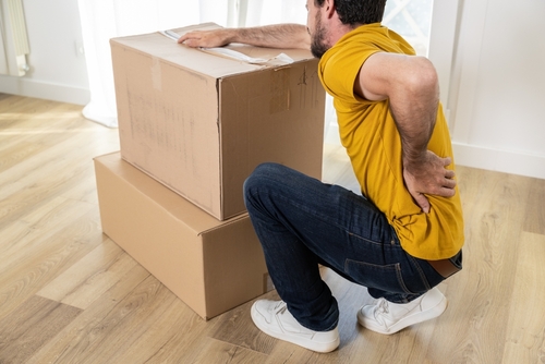 Man crouching beside cardboard boxes and holding his lower back, showing signs of injury while moving or lifting.