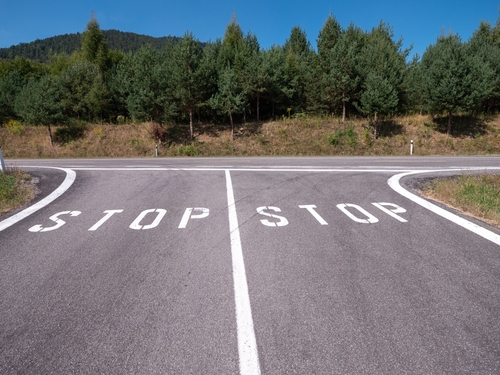 T-junction with two large white "STOP" markings painted on both lanes of the road, surrounded by trees and a blue sky in the background.