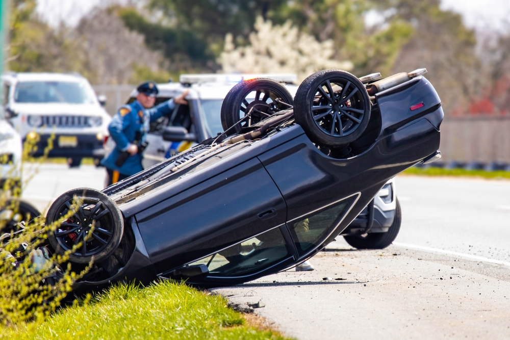 Police officer near an overturned black car after a rollover crash on a highway.